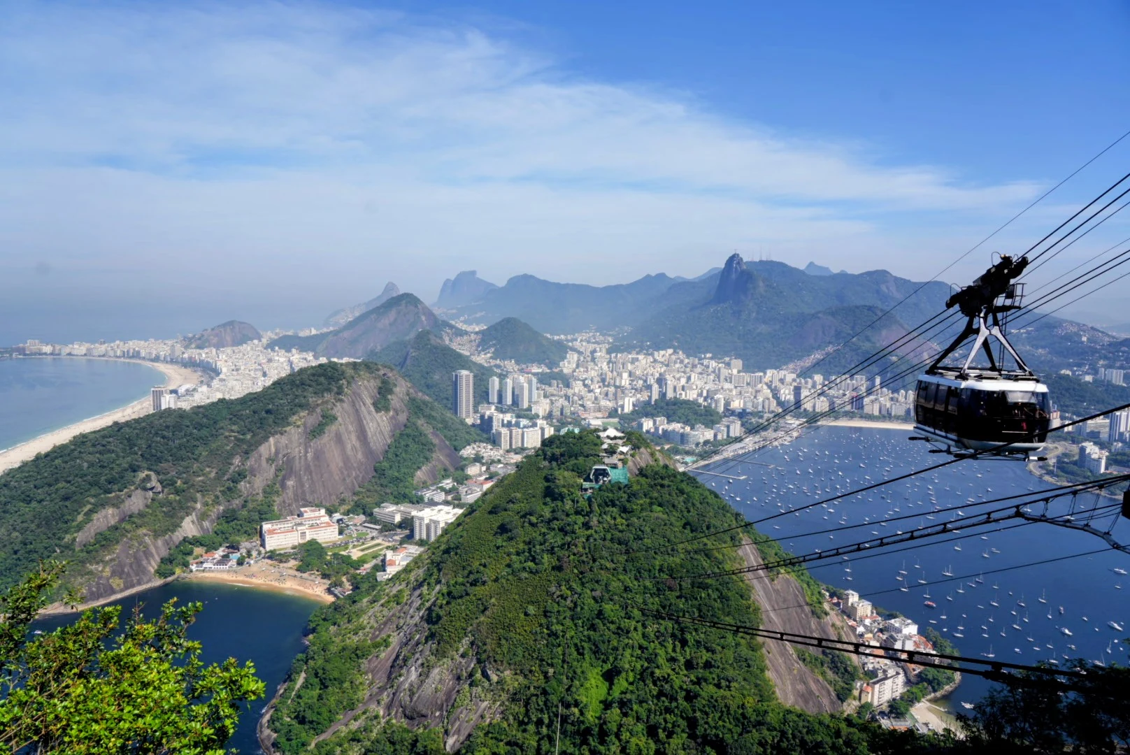 A cable car crossing above Rio de Janeiro, with Copacabana, Botafogo Bay, and Christ the Redeemer in the distance.