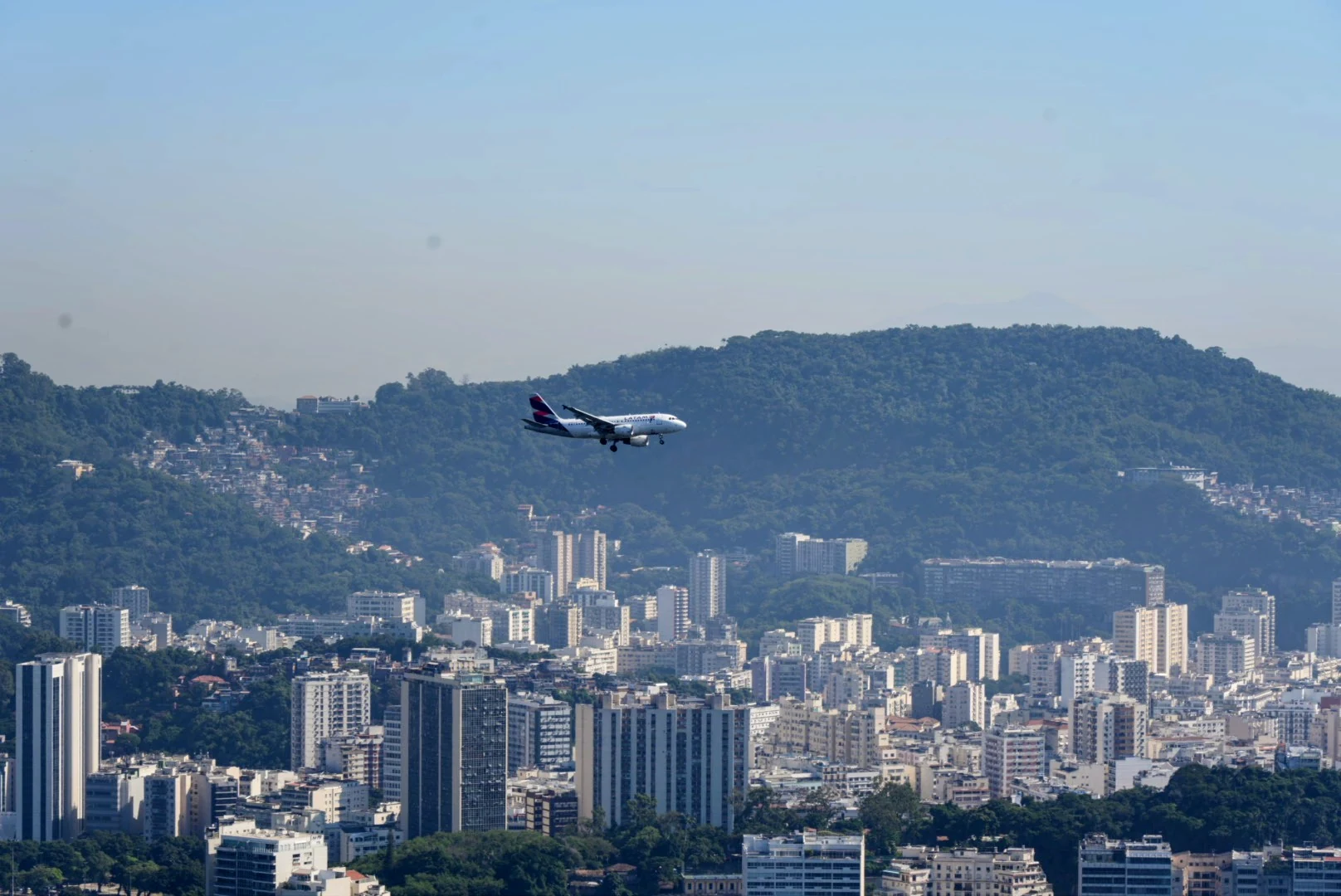 A passenger airplane descending over Rio de Janeiro's high-rises and forested hills.