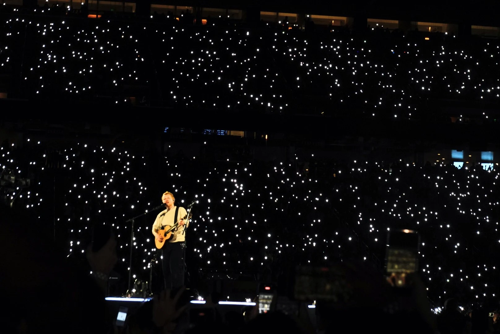 Ed Sheeran performing with an acoustic guitar before a stadium filled with phone lights.