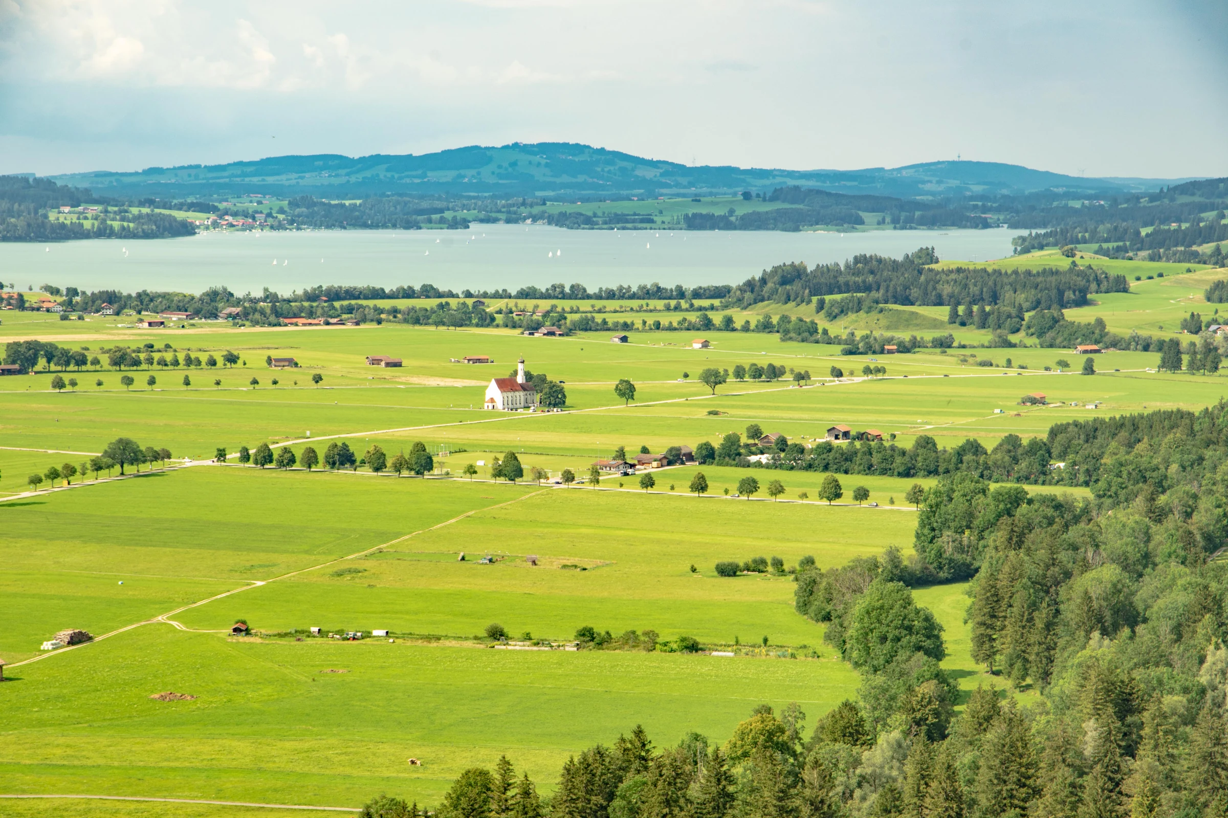 Bright green fields, scattered farmhouses, and a lake across the Bavarian countryside.