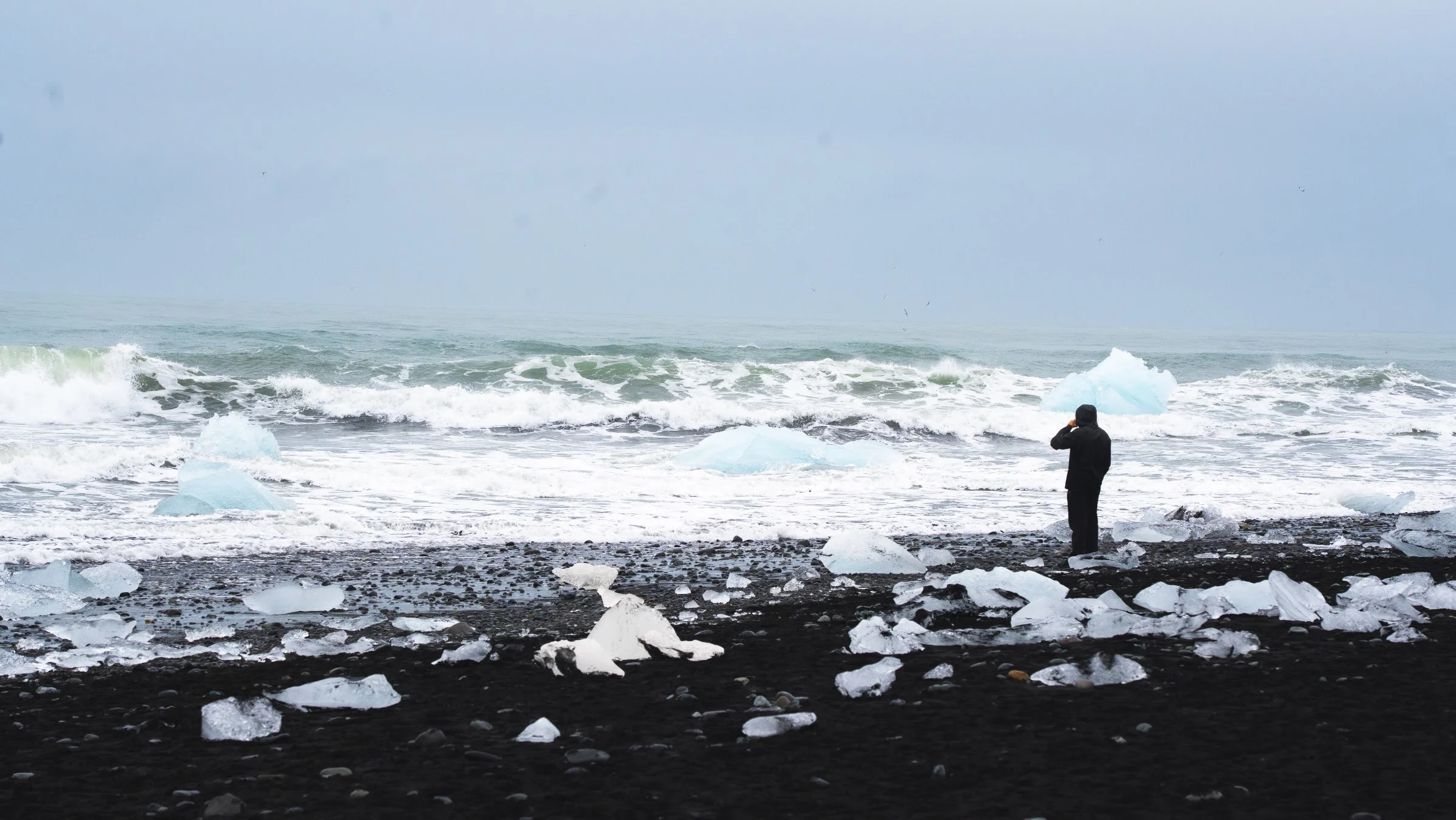 A person watching icy surf break on a black-sand beach.