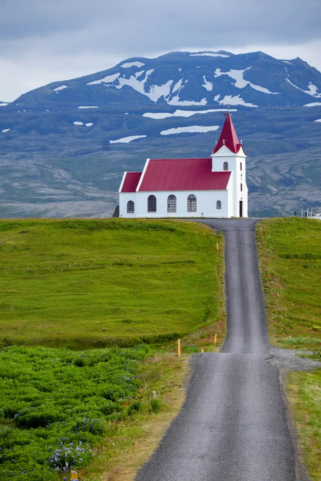 A white church with a red roof at the end of a road beneath a snow-streaked Icelandic mountain.