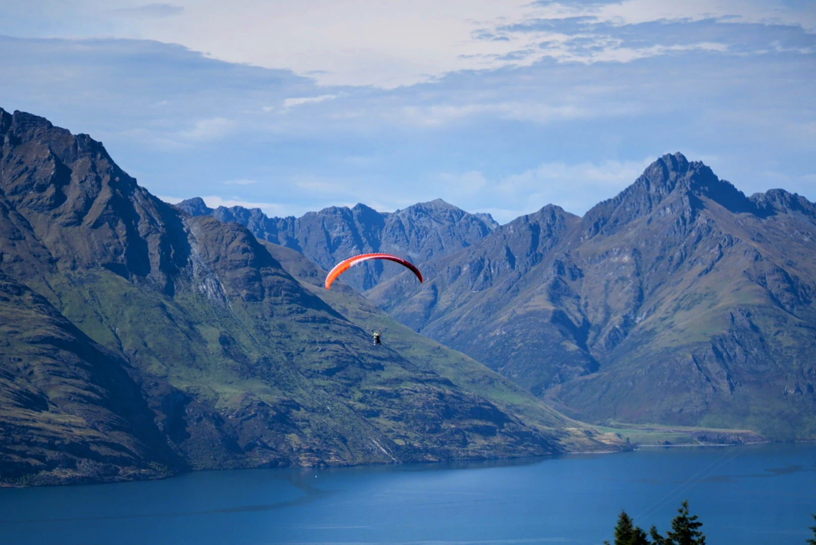 A red paraglider suspended above Lake Wakatipu with rugged mountains beyond.