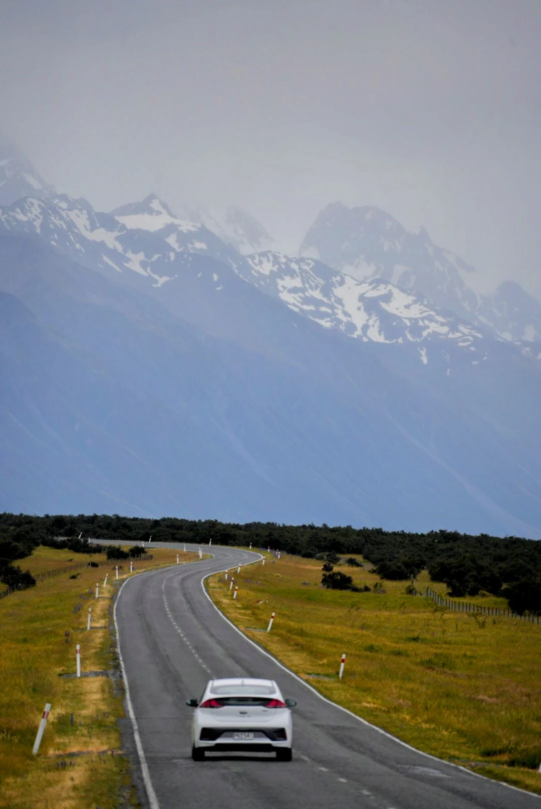 A white car following a winding road toward misty, snow-streaked mountains.