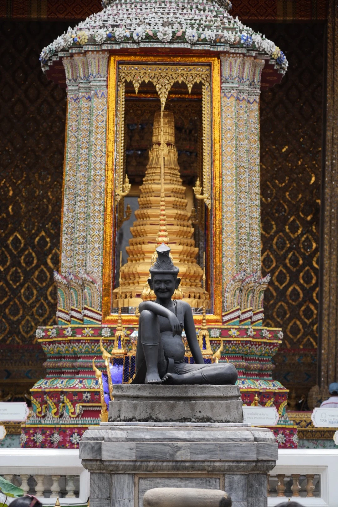 A dark hermit statue aligned with an ornate gilded shrine at the Grand Palace in Bangkok.