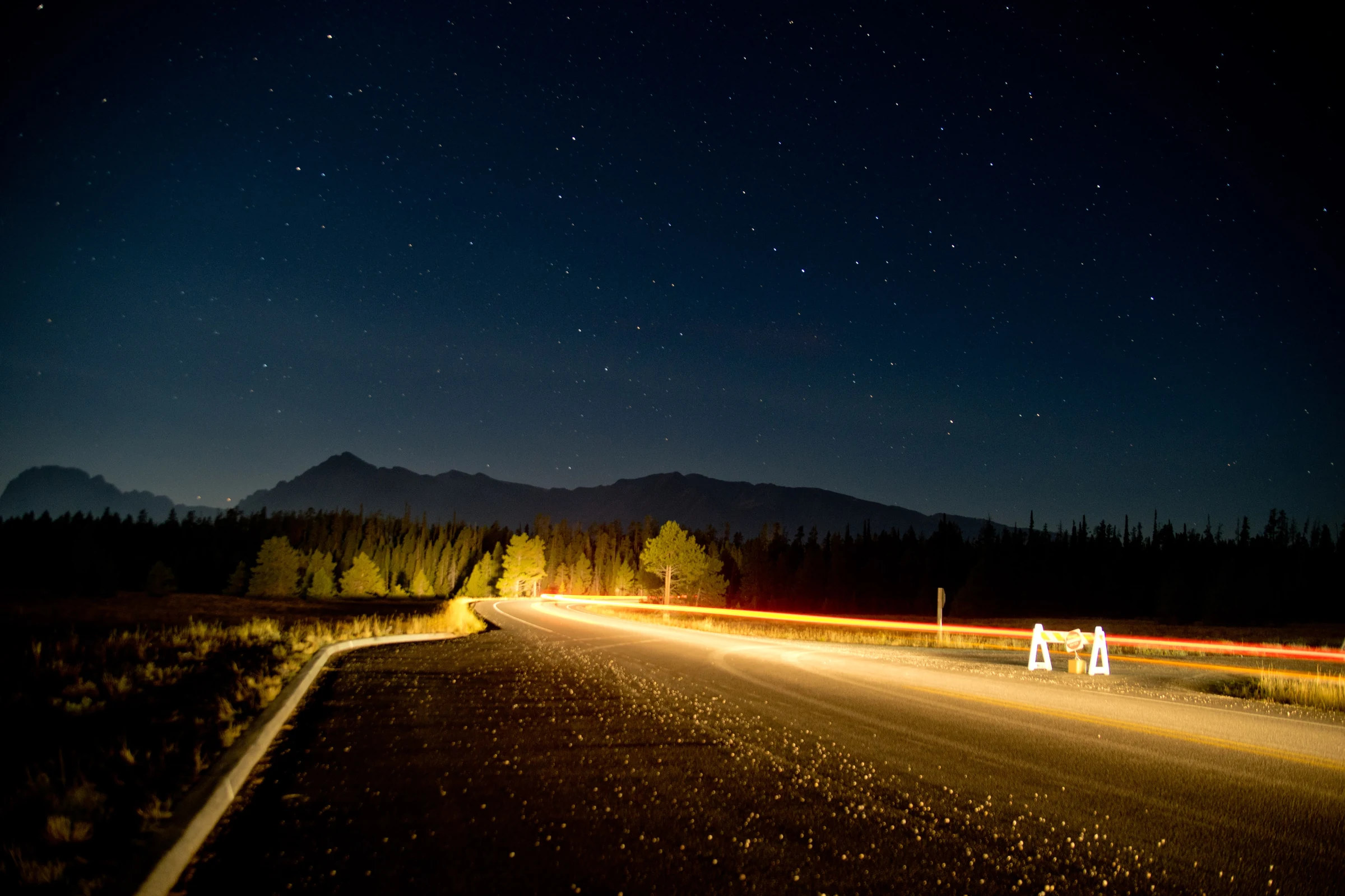 A star-filled night sky above a mountain road traced by car lights.