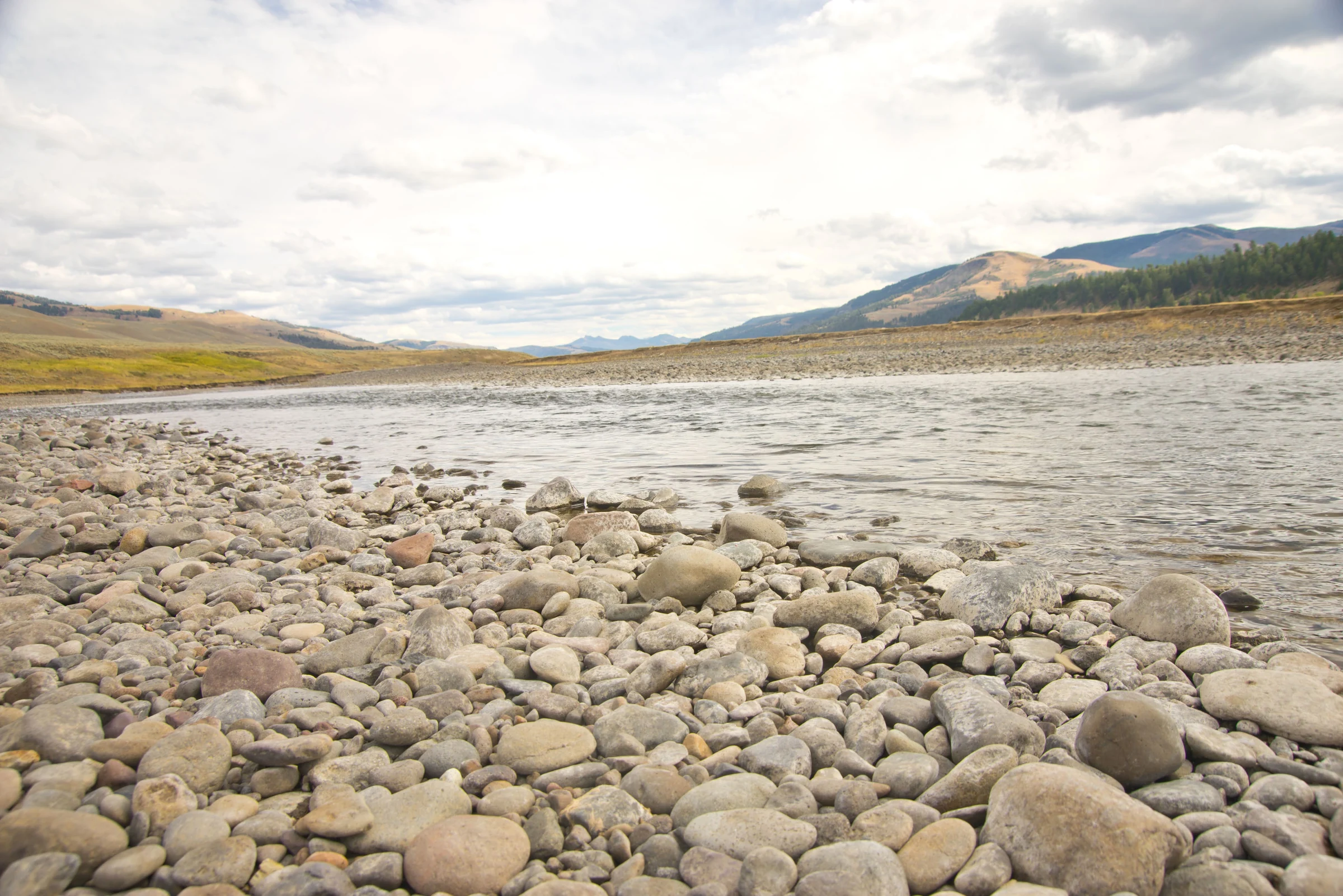 A broad rocky riverbank leading toward mountains beneath a bright sky.